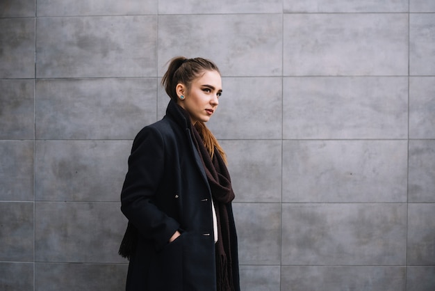 Élégante jeune femme en manteau avec écharpe près du mur gris