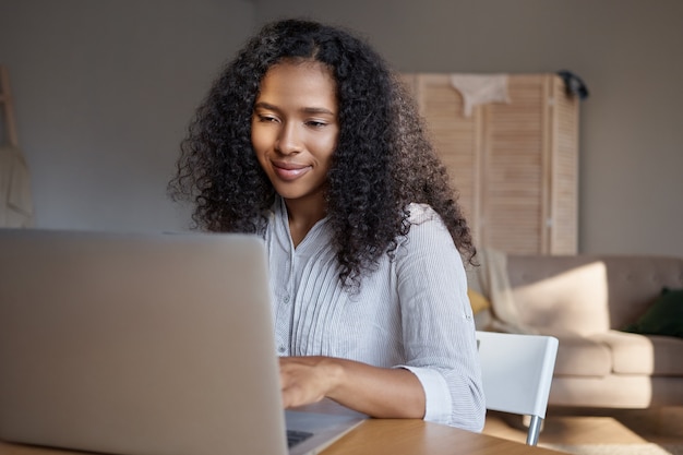 Élégante jeune femme noire aux cheveux bouclés à l'aide d'un ordinateur portable pour le travail à distance. Jolie fille africaine à la recherche d'informations sur internet, bénéficiant d'une connexion sans fil haut débit sur electroni cgadget à la maison