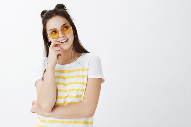 Élégante jeune femme posant avec des lunettes de soleil contre le mur blanc