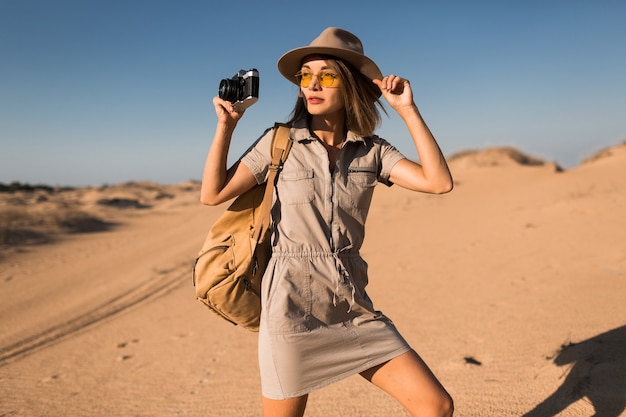 Élégante jeune femme en robe kaki marchant dans le désert, voyageant en Afrique en safari, portant un chapeau et un sac à dos, prenant une photo sur un appareil photo vintage