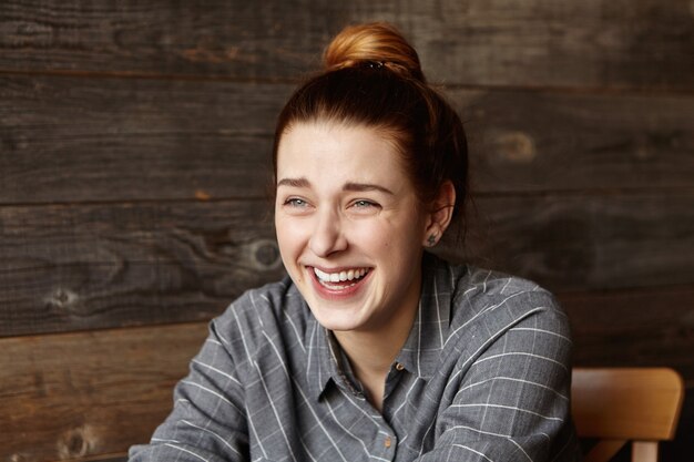 Élégante jeune femme rousse portant chemise à carreaux gris rire aux éclats tout en s'amusant à l'intérieur