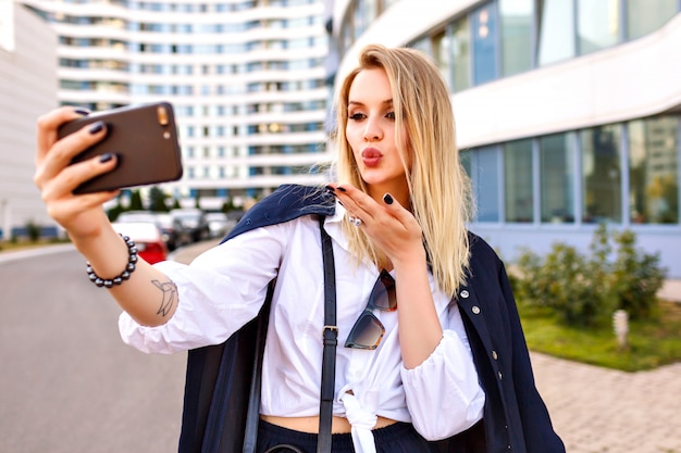 Élégante jeune femme vêtue d'un costume bleu marine à la mode, posant près de bâtiments modernes, d'accessoires à la mode, faisant selfie et vous envoyant de l'air, humeur positive.