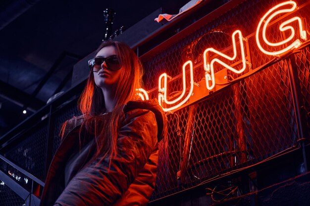 Élégante jeune fille portant un manteau à capuche et des lunettes de soleil debout dans les escaliers d'une discothèque souterraine à l'intérieur industriel