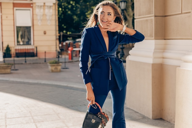 Élégante jolie femme vêtue d'un costume élégant bleu marchant dans la rue tenant le sac à main