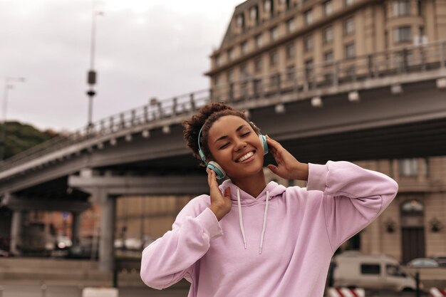 Élégante et joyeuse femme à la peau foncée bouclée en sweat à capuche rose à la mode sourit les yeux fermés et écoute de la musique dans les écouteurs à l'extérieur