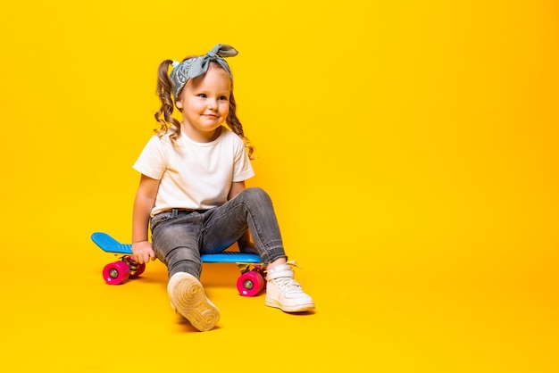 Élégante petite fille enfant fille en tenue décontractée avec planche à roulettes sur mur jaune.