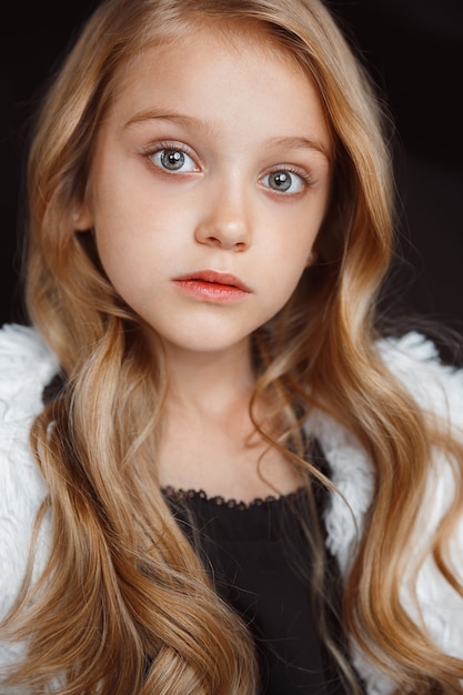 Élégante petite fille posant en tenue blanche isolée sur un mur noir. Belle pose du modèle féminin blonde caucasienne. Émotions humaines, expression faciale, enfance, mode, style.