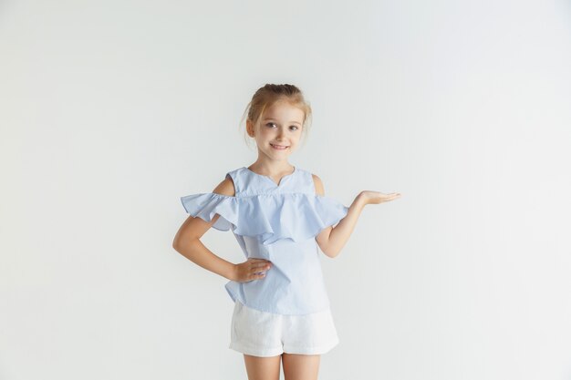 Élégante petite fille souriante posant dans des vêtements décontractés isolés sur fond de studio blanc. Modèle féminin blonde caucasienne. Émotions humaines, expression faciale, enfance. Affichage d'un espace vide, invitant