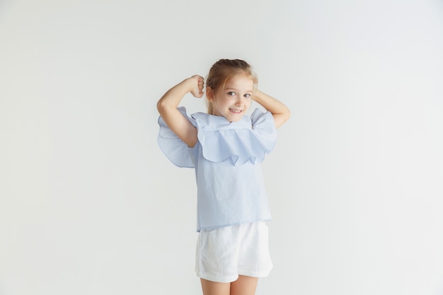 Élégante petite fille souriante posant dans des vêtements décontractés isolés sur fond de studio blanc. Modèle féminin blonde caucasienne. Émotions humaines, expression faciale, enfance. Gagner, célébrer, sourire.