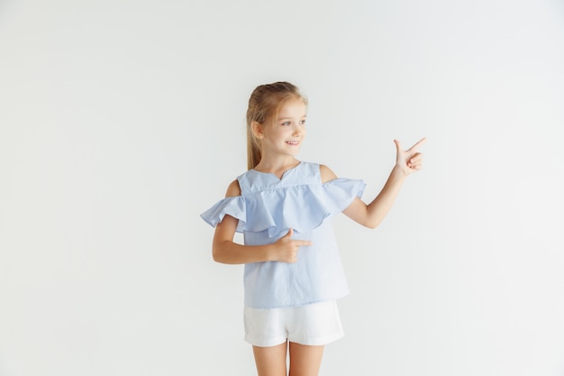 Élégante petite fille souriante posant dans des vêtements décontractés isolés sur fond de studio blanc. Modèle féminin blonde caucasienne. Émotions humaines, expression faciale, enfance. Pointant sur la barre d'espace vide.