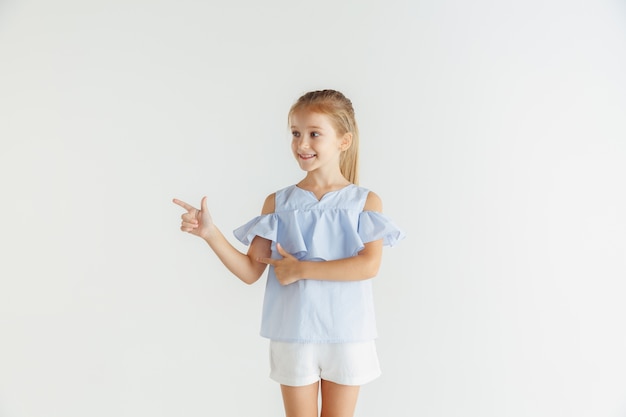 Élégante petite fille souriante posant dans des vêtements décontractés isolés sur un mur blanc. Modèle féminin blonde caucasienne. Émotions humaines, expression faciale, enfance. Pointant sur la barre d'espace vide.