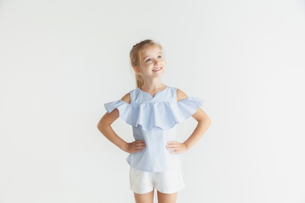 Élégante petite fille souriante posant dans des vêtements décontractés isolés sur un mur blanc. Modèle féminin blonde caucasienne. Émotions humaines, expression faciale, enfance. Souriant, tenant les mains sur les hanches.