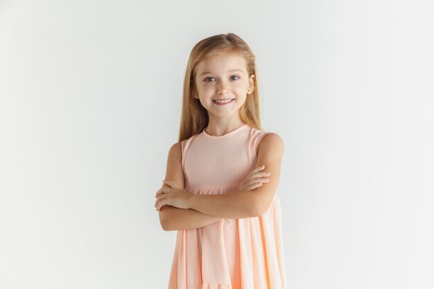 Élégante petite fille souriante qui pose en robe isolée sur fond de studio blanc. Modèle féminin blonde caucasienne. Émotions humaines, expression faciale, enfance. Debout avec les mains croisées.