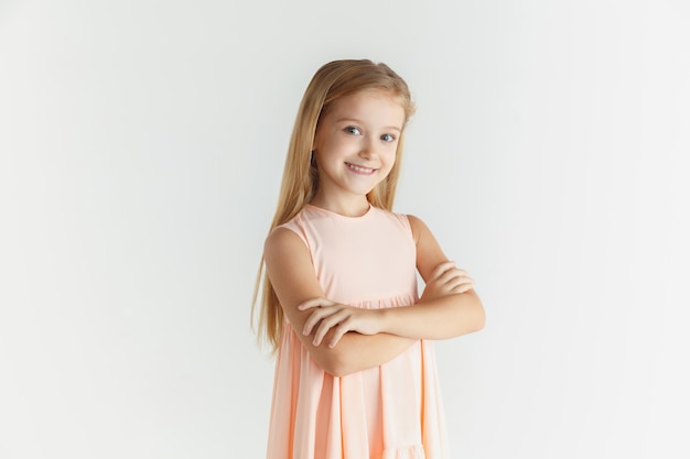 Élégante petite fille souriante qui pose en robe isolée sur fond de studio blanc. Modèle féminin blonde caucasienne. Émotions humaines, expression faciale, enfance. Debout avec les mains croisées.