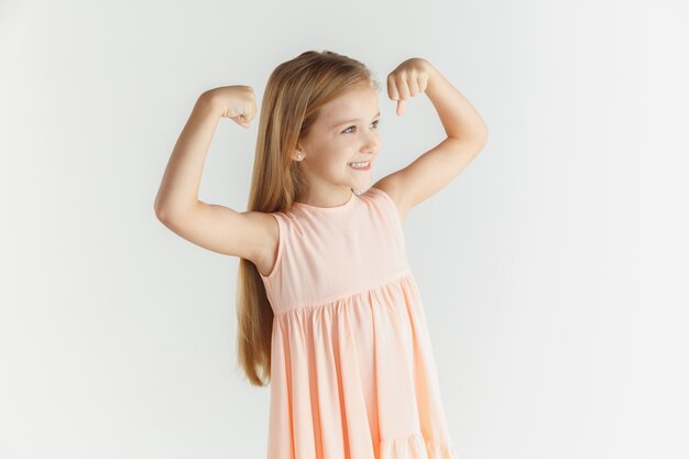 Élégante petite fille souriante qui pose en robe isolée sur fond de studio blanc. Modèle féminin blonde caucasienne. Émotions humaines, expression faciale, enfance. Gagner, célébrer, a l'air heureux.