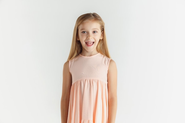 Élégante petite fille souriante qui pose en robe isolée sur fond de studio blanc. Modèle féminin blonde caucasienne. Émotions humaines, expression faciale, enfance. Se demanda, regardant la caméra.