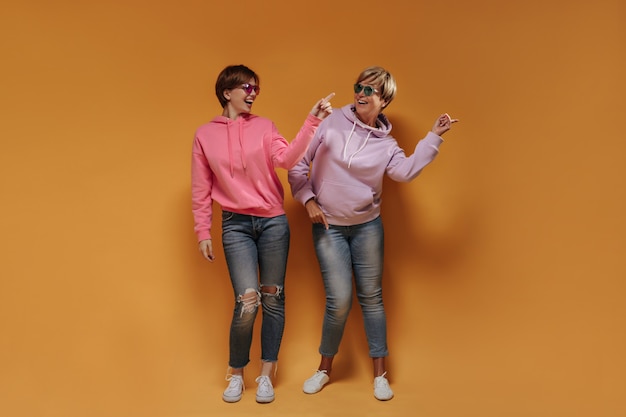 Élégantes dames aux cheveux courts dans des lunettes lumineuses, de larges sweats à capuche, des baskets blanches et des jeans skinny dansant et souriant sur fond orange.