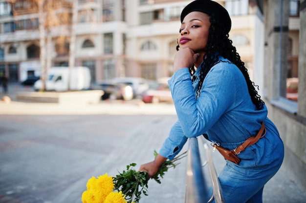 Élégantes femmes afro-américaines à la mode en jeans et béret noir avec bouquet de fleurs jaunes posé en plein air par temps ensoleillé contre un bâtiment moderne bleu