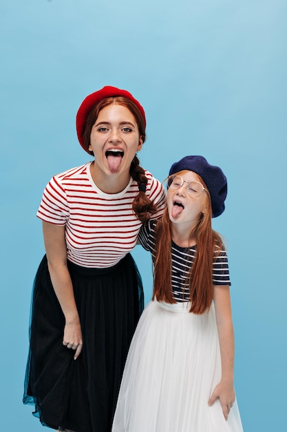 Élégantes jeunes femmes aux cheveux rouges dans des bérets brillants, des jupes fraîches et des t-shirts élégants montrant des langues sur fond bleu isolé