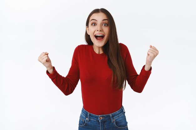 Élevée et inspirée, excitée fille brune en pull rouge, poings serrés en triomphe, ayant des aspirations souriantes étonnées et étonnées, voir l'équipe gagner, atteindre un objectif ou réussir, fond blanc