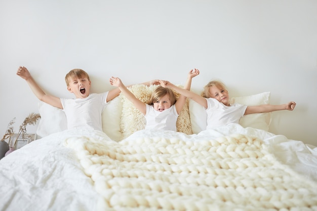 Lever et briller. Tir horizontal isolé de trois frères et sœurs petite sœur et ses deux frères aînés portant des t-shirts blancs identiques assis sur le lit, étirant les bras et bâillant le matin