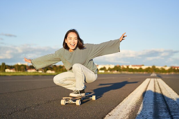 Liberté et bonheur Heureuse fille asiatique chevauchant son longboard sur une route ensoleillée vide riant et souriant faisant du skateboard