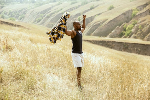 Liberté. Jeune voyageur afro-américain debout confiant dans les prés verts en journée d'été ensoleillée. Ça a l'air sérieux, a levé les mains. L'été, les vacances, les vacances, le repos.