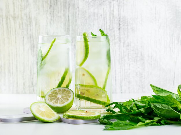 Limonade dans des verres au citron, basilic, planche à découper vue latérale sur blanc et grungy