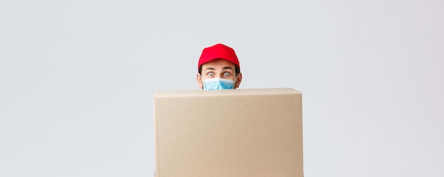 Photo gratuite livraison de colis et de colis covid19 commandes de quarantaine et de transfert courrier mignon maladroit premier jour au travail se cachant derrière une grande boîte porter un masque facial et une casquette uniforme rouge fond gris