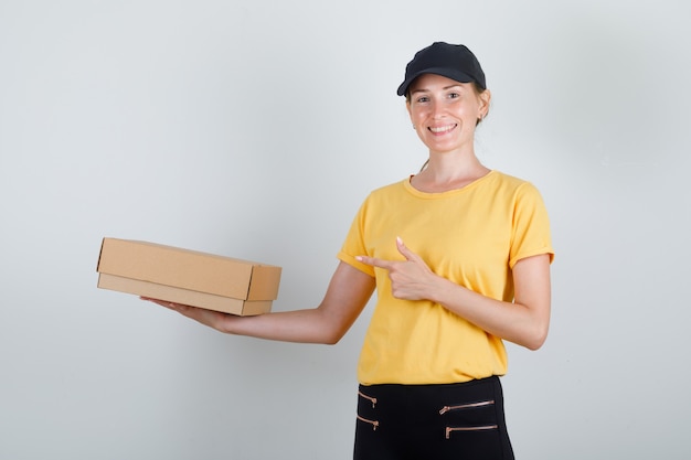 Livraison femme doigt pointé sur boîte en carton en t-shirt, pantalon, casquette et à heureux