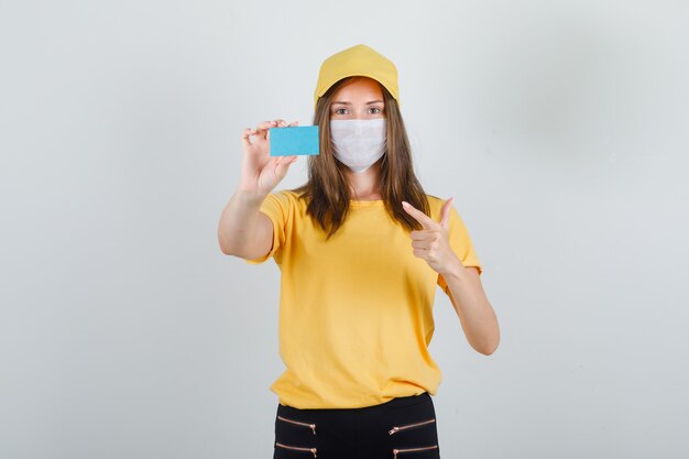 Livraison femme doigt pointé sur carte bleue en t-shirt, pantalon, casquette et masque et à la joie