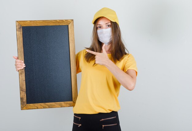 Livraison femme doigt pointé sur tableau noir en t-shirt, pantalon, casquette et masque