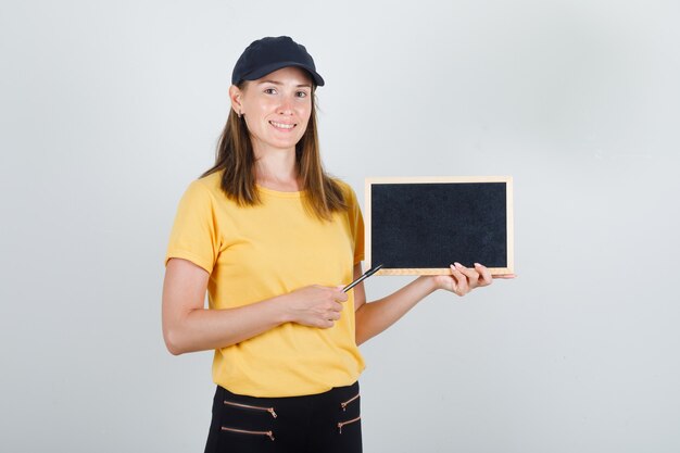 Livraison femme pointant le stylo au tableau noir en t-shirt, pantalon, casquette et à la joie.