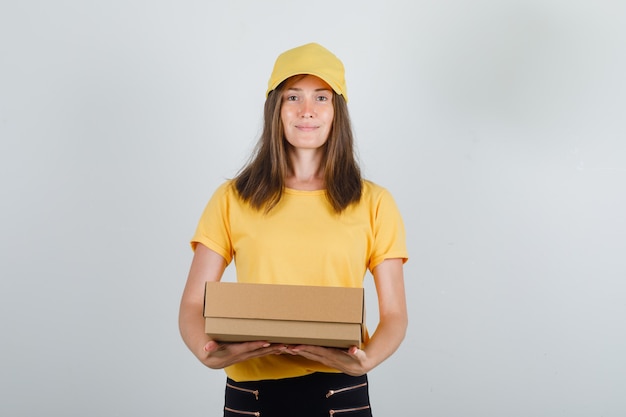 Livraison femme tenant une boîte en carton et souriant en t-shirt jaune, pantalon et casquette