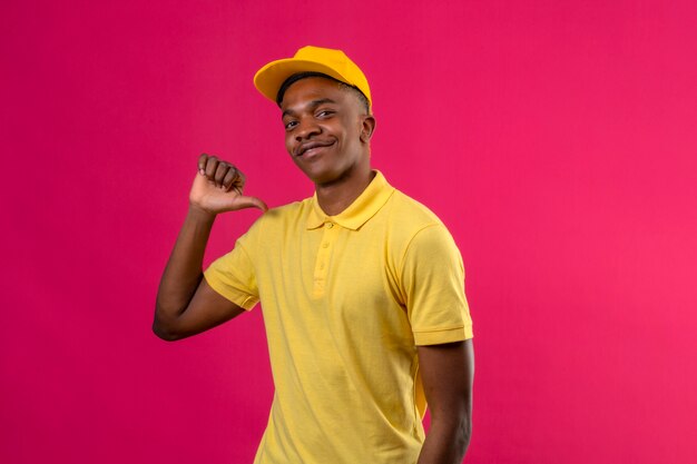 Livraison homme afro-américain en polo jaune et casquette à la confiance en pointant avec le pouce à lui-même fier debout sur rose