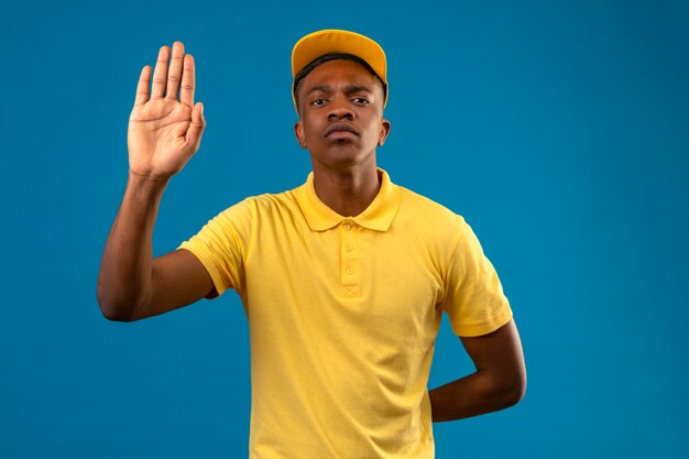 Livraison homme afro-américain en polo jaune et casquette debout avec la main ouverte faisant panneau d'arrêt avec un geste de défense d'expression sérieuse et confiante sur bleu isolé