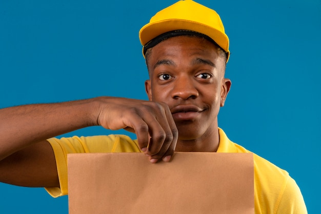 Photo gratuite livraison homme afro-américain en polo jaune et casquette holding paper package smiling friendly sur bleu isolé