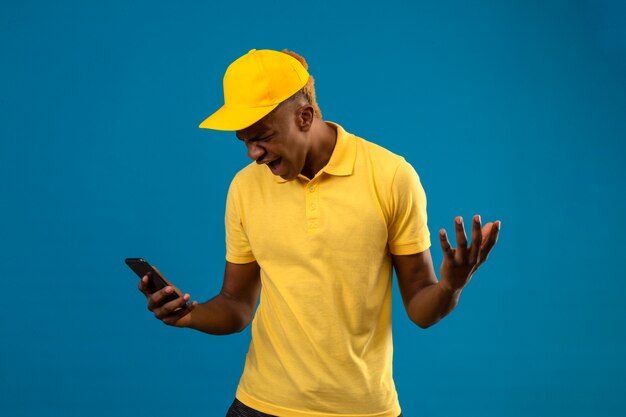 Livraison homme afro-américain en polo jaune et casquette regardant téléphone mobile avec expression de colère levant les mains frustré debout sur bleu isolé