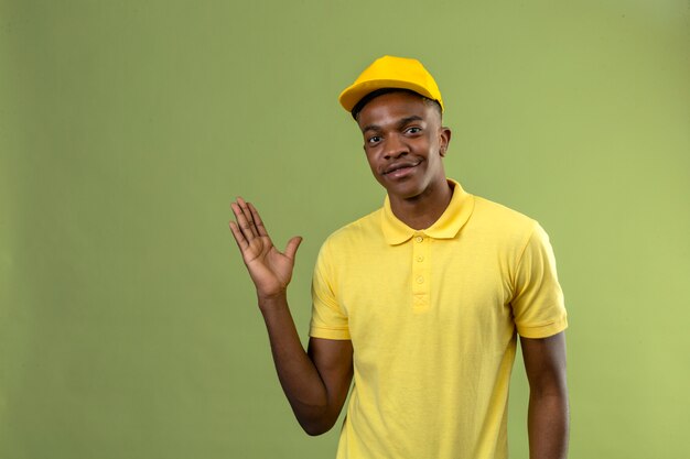 Livraison homme afro-américain en polo jaune et casquette souriant amical avec main levée debout sur le vert