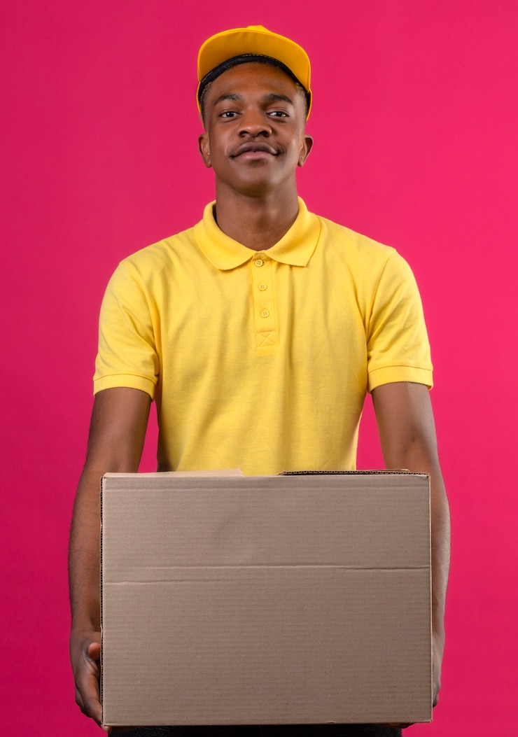 Photo gratuit livraison homme afro-américain en polo jaune et casquette tenant une boîte en carton à la confiance debout sur rose