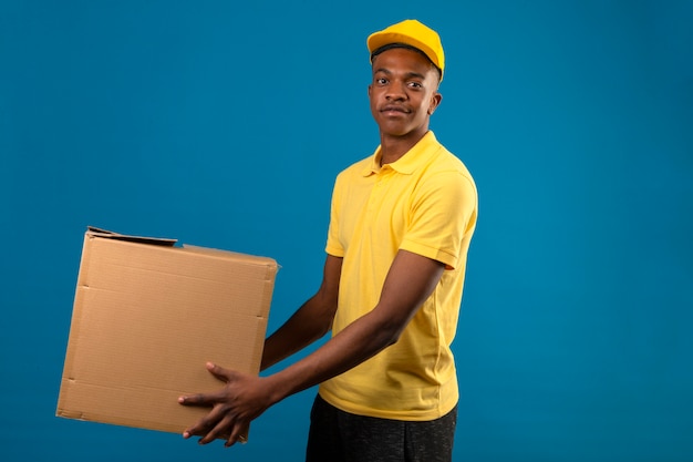 Livraison homme afro-américain en polo jaune et casquette tenant des boîtes en carton avec sourire amical debout sur bleu isolé