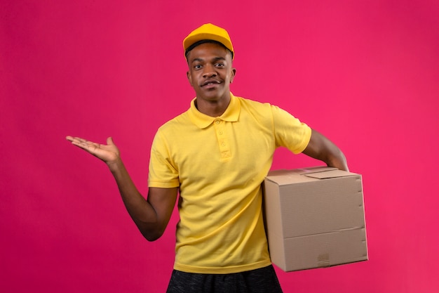 Livraison homme afro-américain en polo jaune et casquette tenant le paquet de boîte présentant et pointant avec la paume de la main debout sur rose isolé