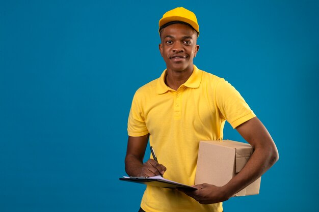 Livraison homme afro-américain en polo jaune et casquette tenant le presse-papiers et boîte en carton avec sourire confiant debout sur bleu isolé