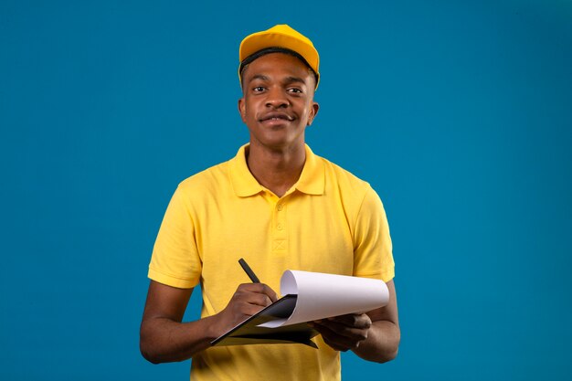 Livraison homme afro-américain en polo jaune et casquette tenant le presse-papiers écrit quelque chose à la confiance et fier debout sur bleu isolé