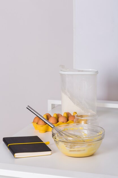 Livre de recettes; carton d&#39;oeufs; farine et oeufs fouettés sur une table blanche contre un mur blanc