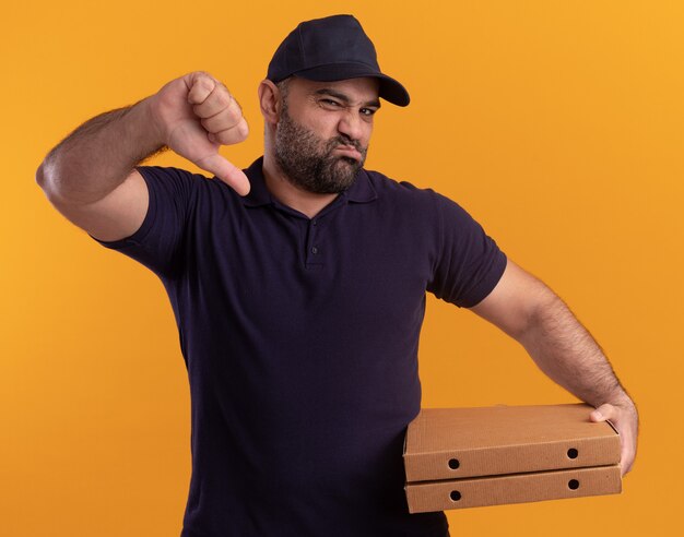 Livreur d'âge moyen mécontent en uniforme et casquette tenant des boîtes à pizza montrant le pouce vers le bas isolé sur un mur jaune