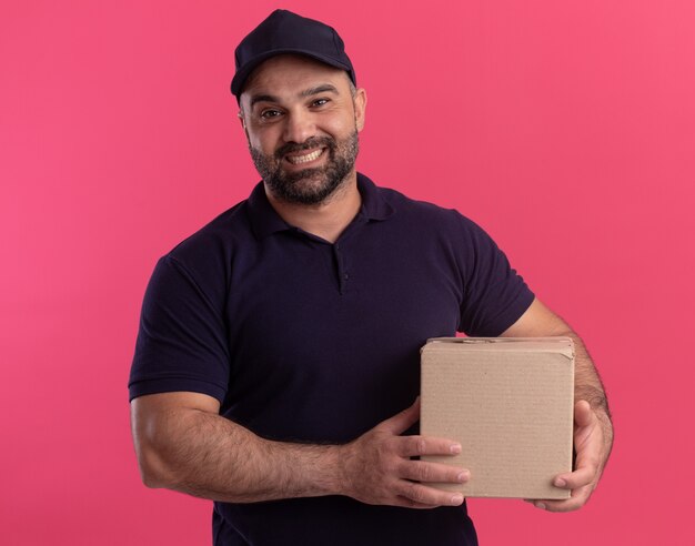 Livreur d'âge moyen souriant en uniforme et casquette tenant une boîte isolée sur un mur rose