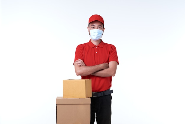 Livreur asiatique portant un masque facial en uniforme rouge et boîte à colis sur blanc.