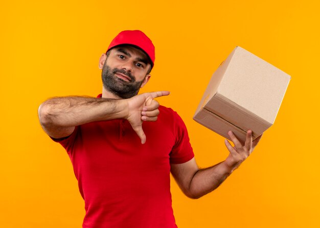 Livreur barbu en uniforme rouge et cap holding box package montrant les pouces vers le bas debout sur le mur orange