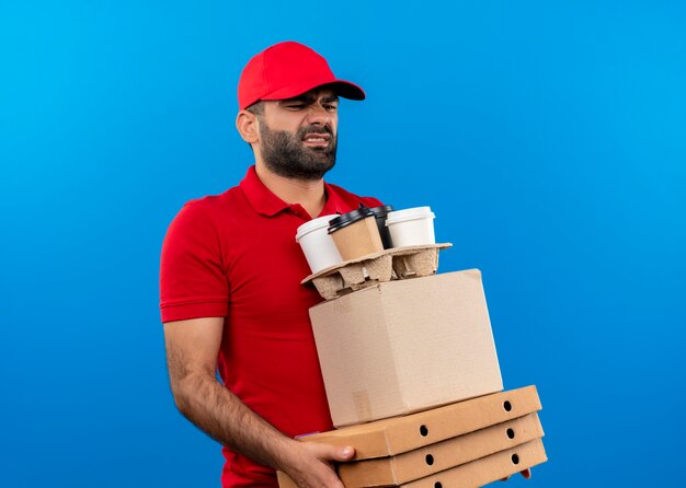 Livreur barbu en uniforme rouge et cap tenant des boîtes en carton à côté mécontent de fatigue et surmené debout sur mur bleu